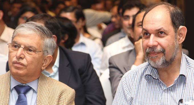 Hormusji N. Cama, at right in a checked shirt, seated in the audience at an industry gathering beside a senior colleague in a jacket and tie, with further delegates behind them.