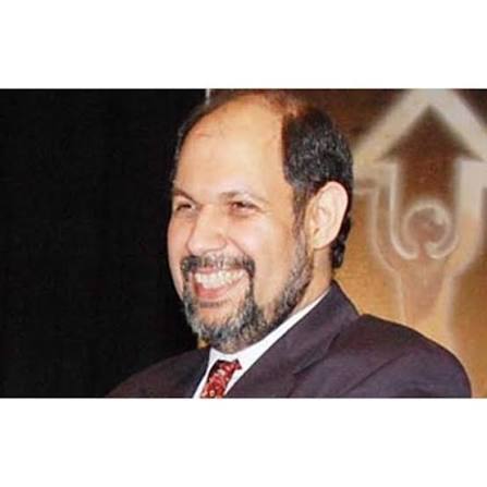 Hormusji N. Cama smiling broadly in a dark suit and patterned tie at a public event, photographed against a dark backdrop bearing a pale emblem.